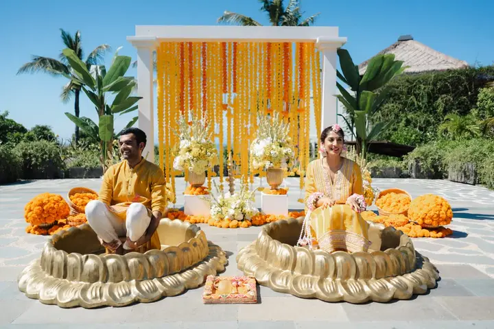 A bride and groom in yellow seated for their haldi before a backdrop of marigolds