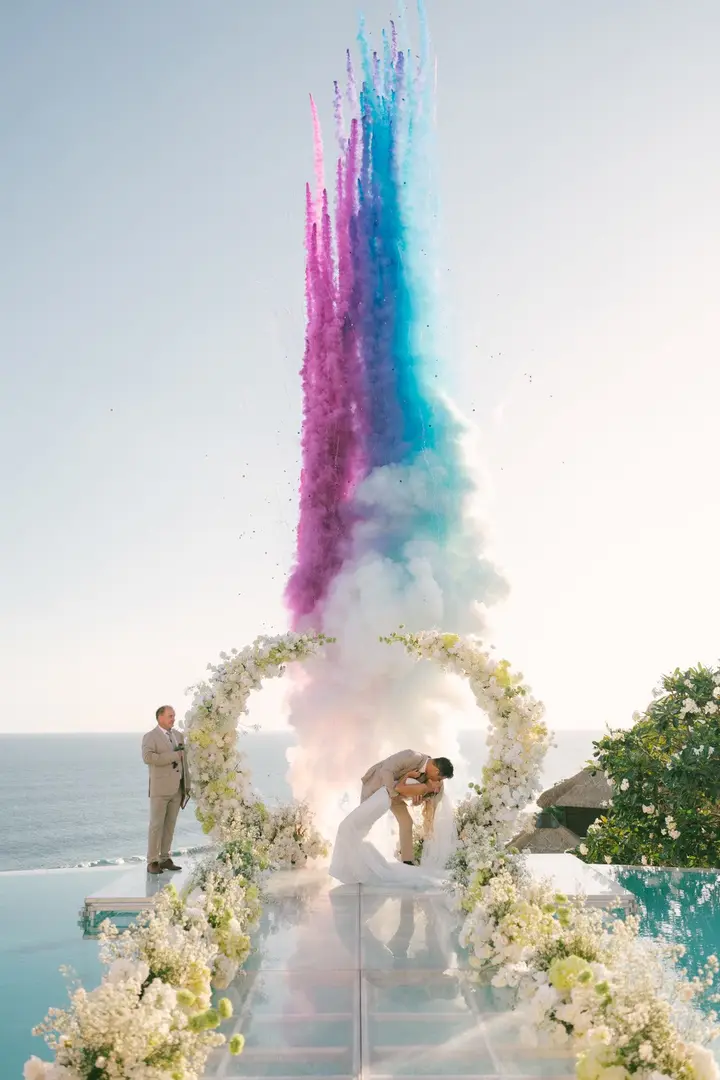Pink and blue smoke rising above a floral ceremony arch on a clifftop
