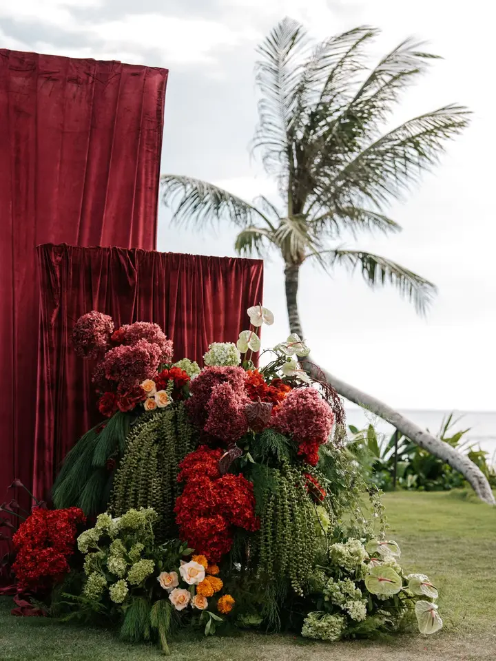 A towering installation of red and green florals before red velvet drapes, with palms and the ocean behind
