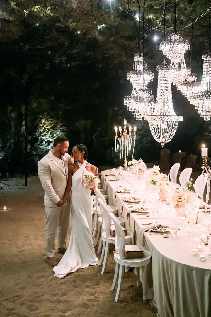 Natalie and Max beside their long reception table beneath crystal chandeliers at night
