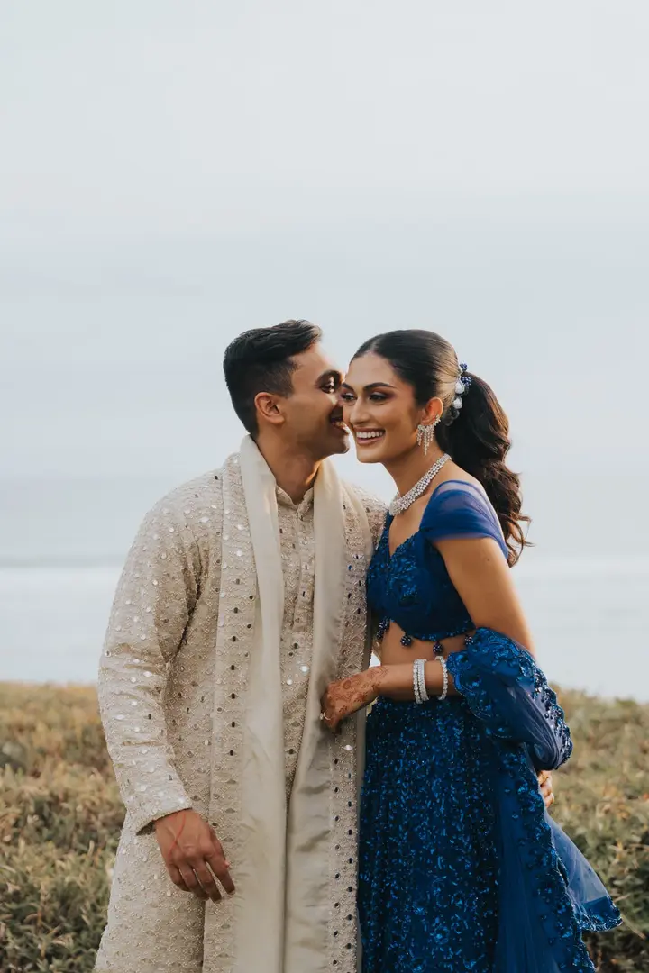 A couple in a deep blue lehenga and an ivory sherwani, smiling together outdoors beneath a soft sky