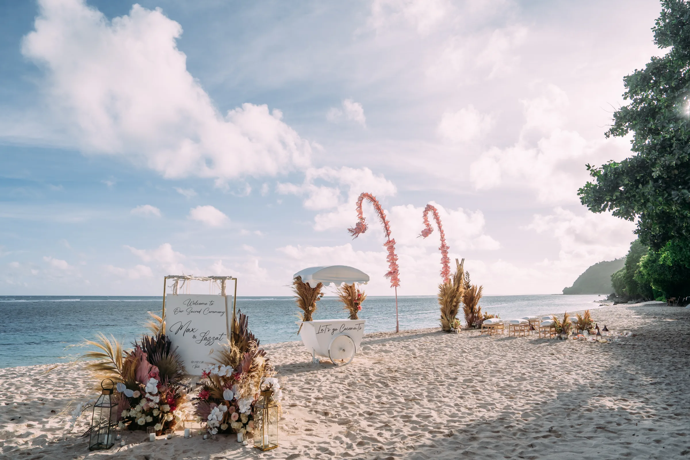 A beachside ceremony setup with pampas grass arches, lounge seating, and a welcome sign by the ocean