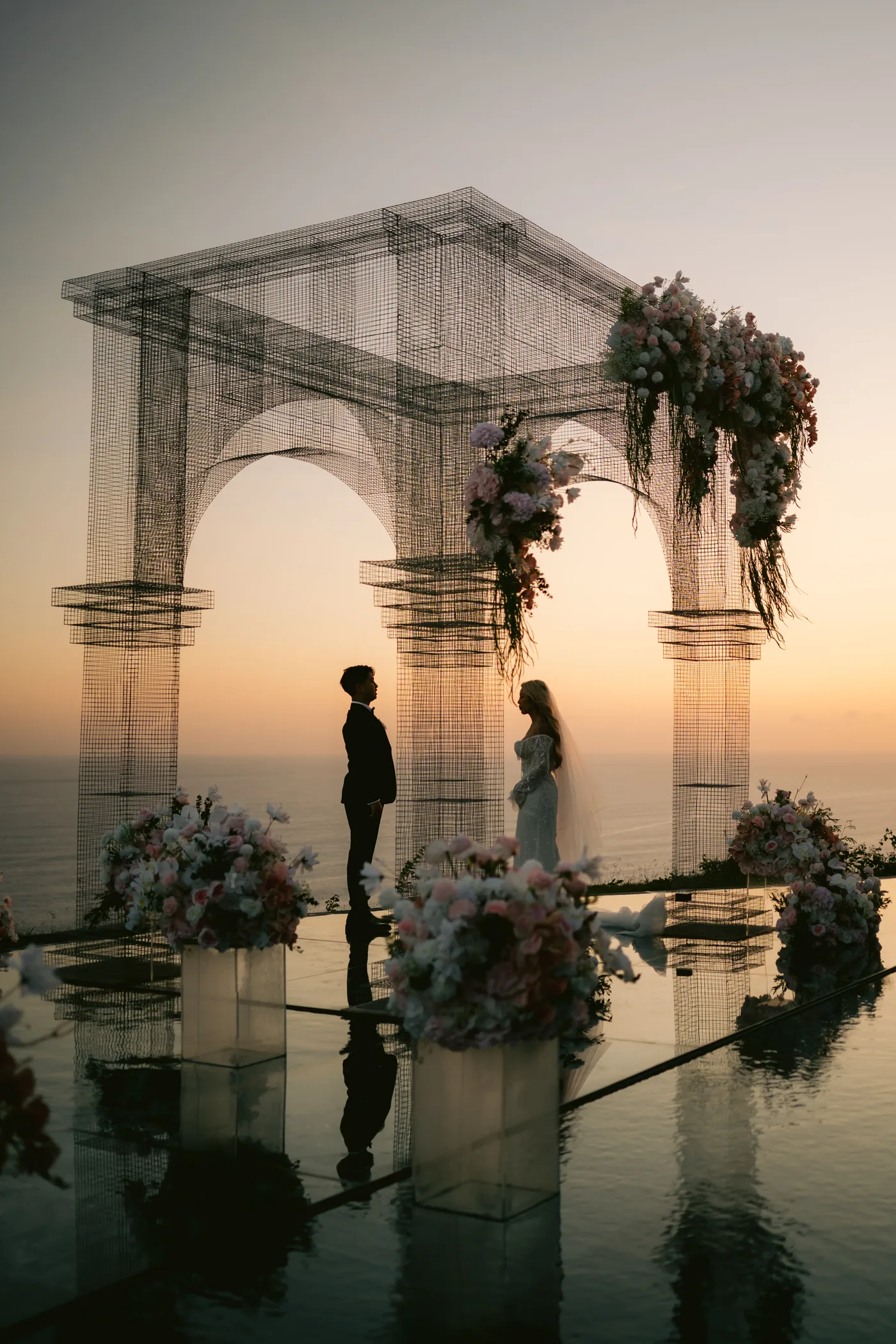 Bride and groom face each other in silhouette beneath a wire-frame arch at sunset
