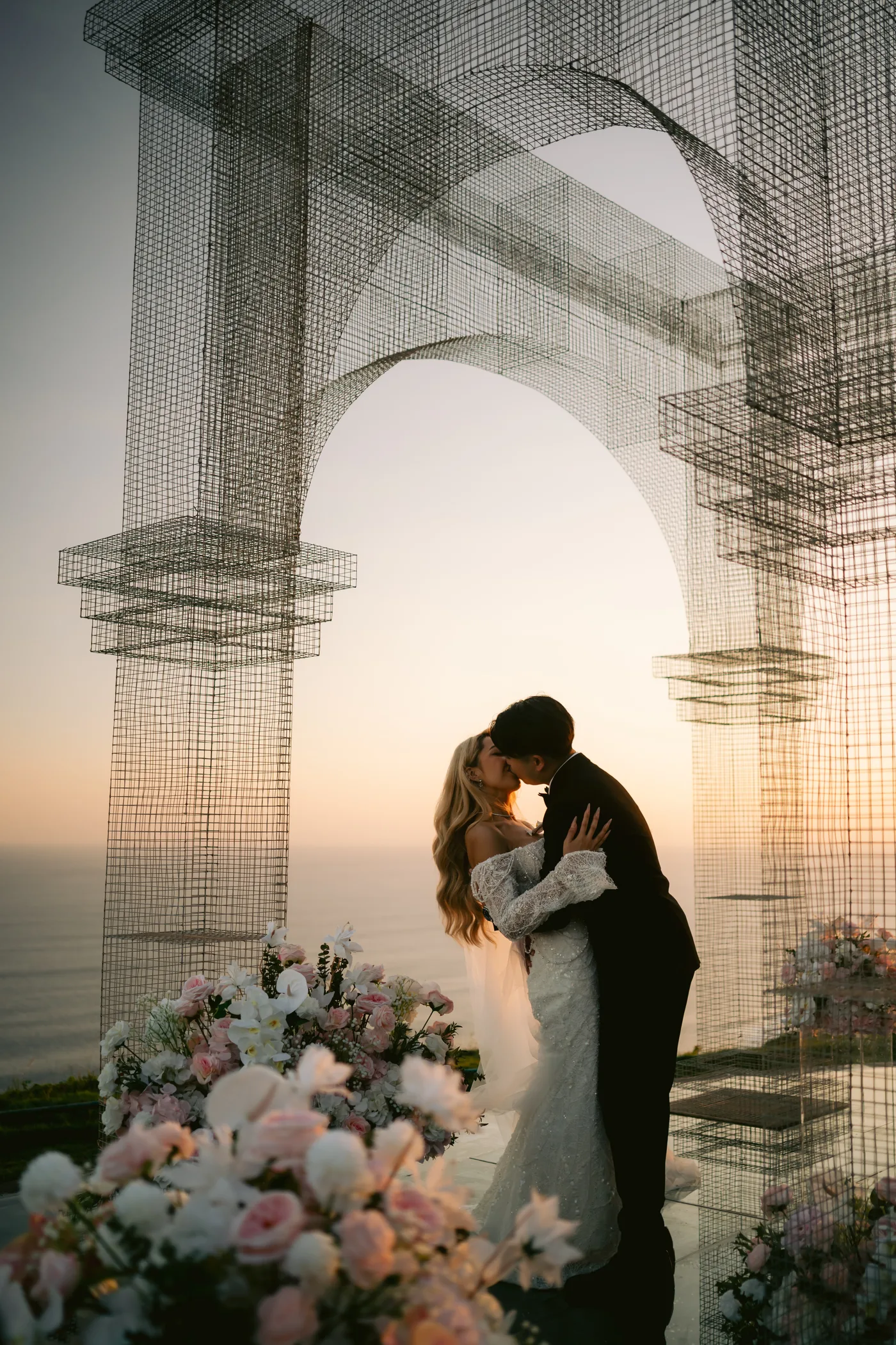 The couple kiss beneath the wire-frame arch as the sun sets over the ocean