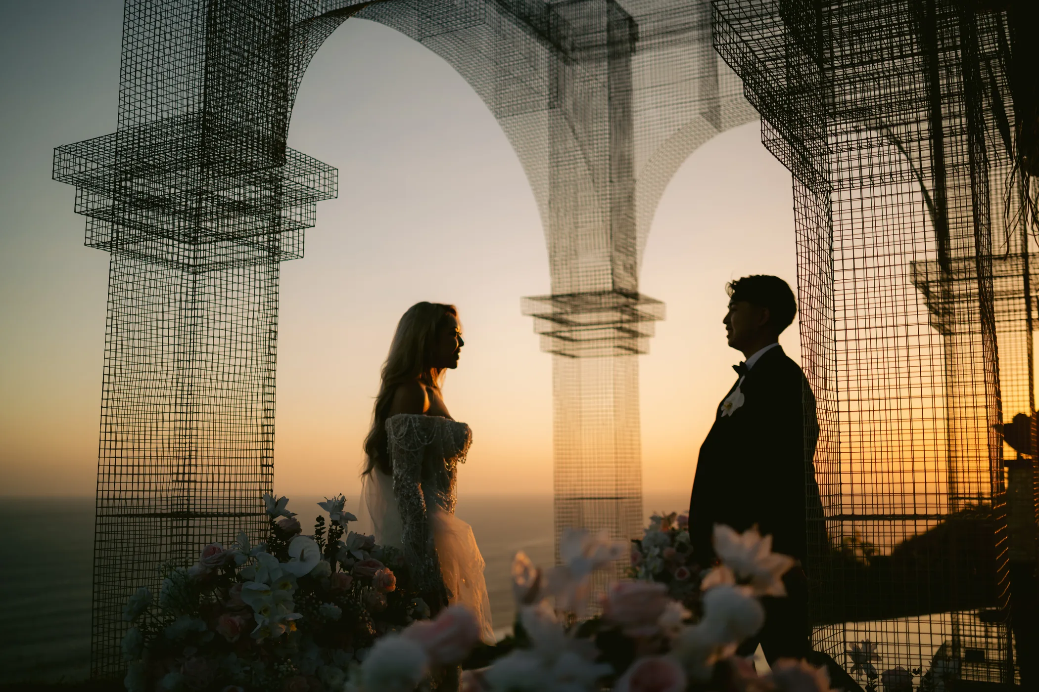 The couple beneath the wire-frame arch, silhouetted against a warm sunset sky