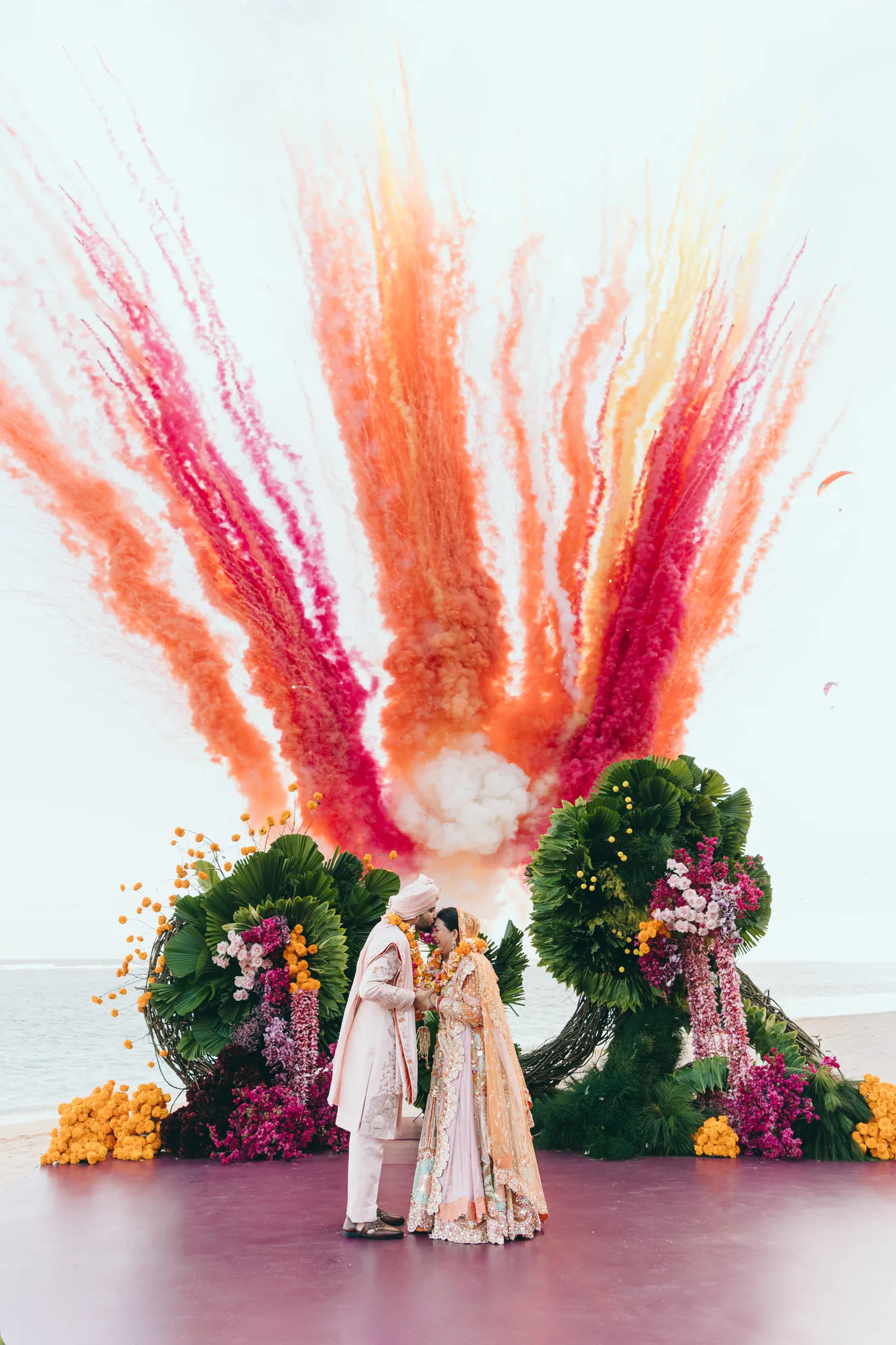 Bride and groom embrace beneath a burst of orange and pink ceremonial smoke