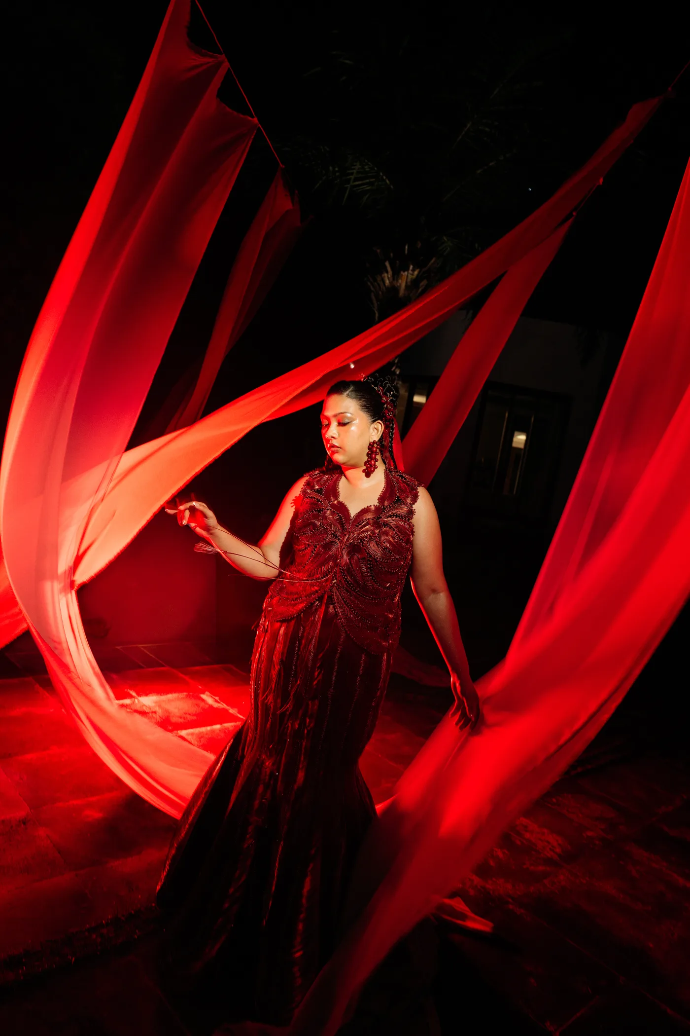The bride in a beaded red gown amid flowing red silk drapes at night